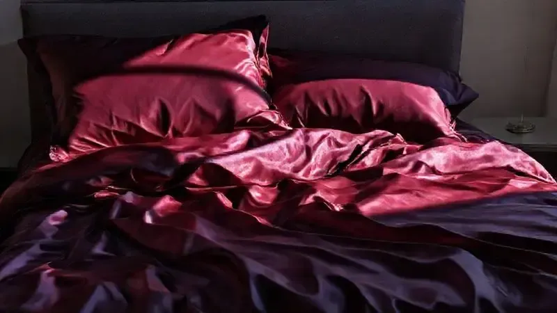 Cama de casal luxuosa com lençóis de seda ou cetim em cor escura, destacando o brilho e a sofisticação.