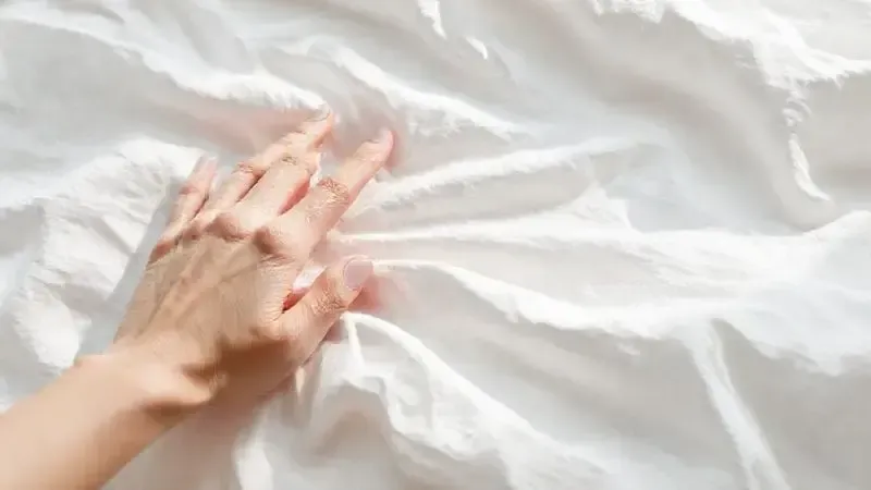 Ilustração de close-up de uma mão tocando um lençol 100% algodão branco, destacando a textura macia e o conforto do tecido.